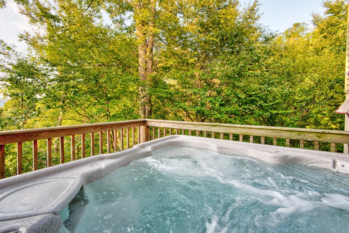 A serene outdoor covered deck housing a Bullfrog Spas hot tub, surrounded by lush greenery and a wooden railing. The scene suggests relaxation, with the hot tub invitingly set against a natural backdrop, ideal for soaking after adventures or during downtime.