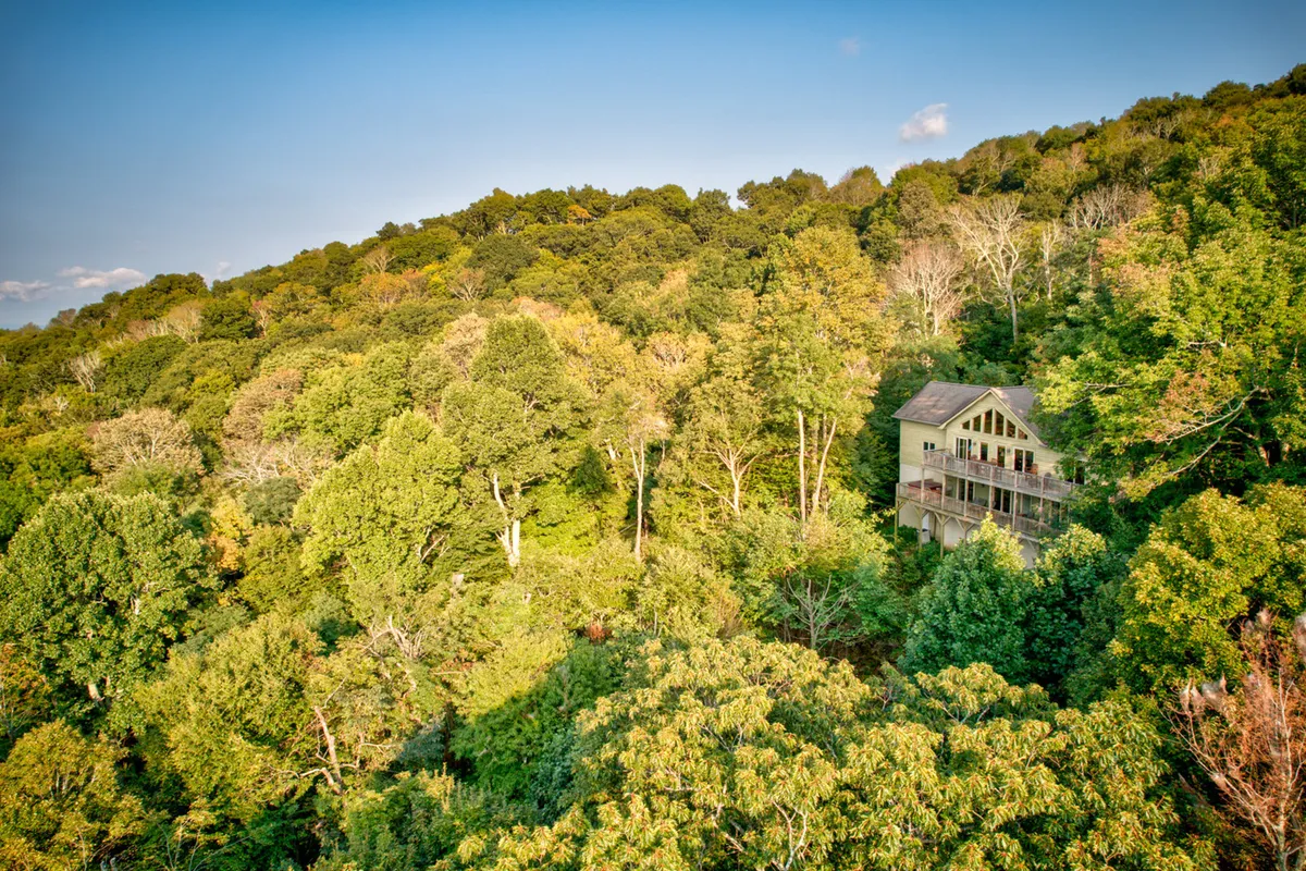 This image presents the home nestled among lush green trees with distant hills visible in the background. The setting feels secluded and immersed in nature, with the deck and surrounding foliage inviting outdoor exploration and relaxation.