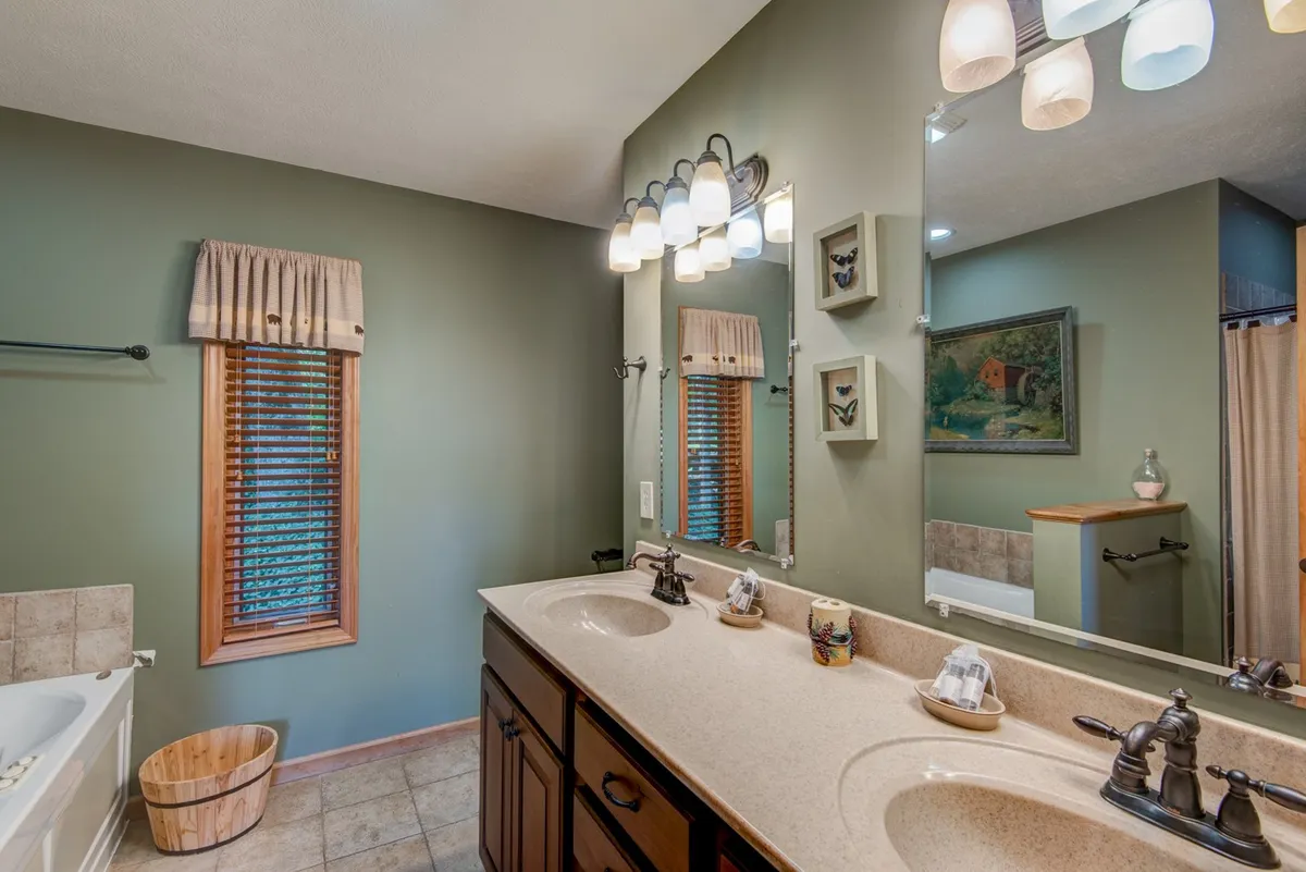 A calm and serene bathroom featuring a double vanity sink with a framed mirror, tasteful wall artwork, and a window with blinds. The atmosphere is designed for comfort and peaceful self-care routines.