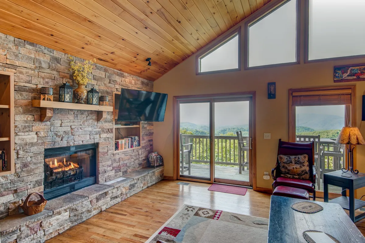 A cozy living room with a central stone fireplace ablaze, large windows framing mountain views, a wooden ceiling, and decorative shelving. The seating is arranged around the fireplace, creating a warm, inviting atmosphere.