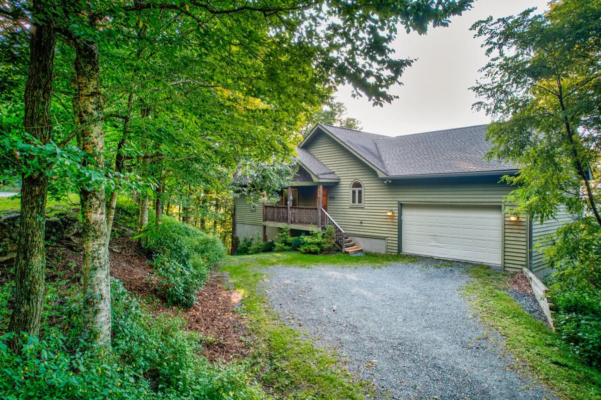 This image displays the exterior of the house nestled between lush greenery, with a clear view of the driveway and welcoming porch. The surrounding landscape hints at nearby hiking paths and the tranquil mountain environment ideal for outdoor adventures.