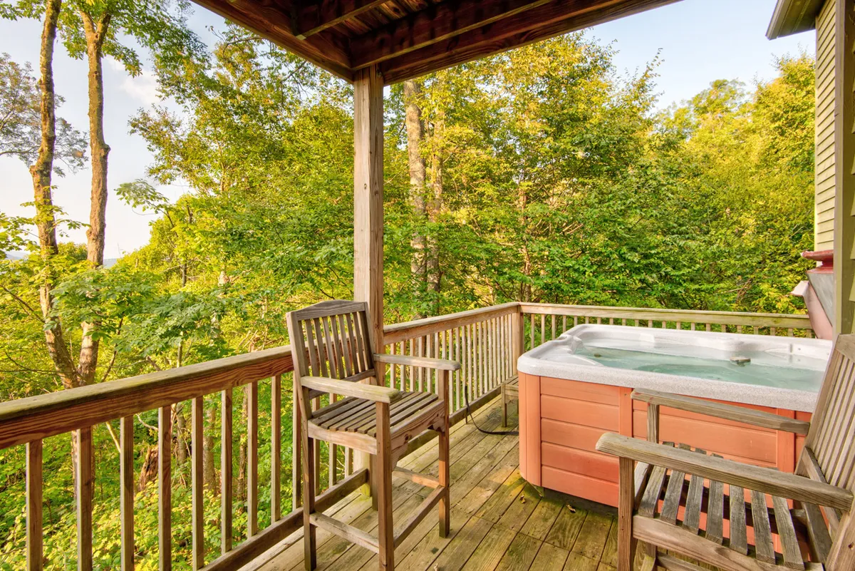 A spacious wooden deck featuring a bubbling hot tub surrounded by trees, with bar stools and seating. The setting invites relaxation under the open sky, perfect for stargazing or unwinding after a day of exploring local attractions.