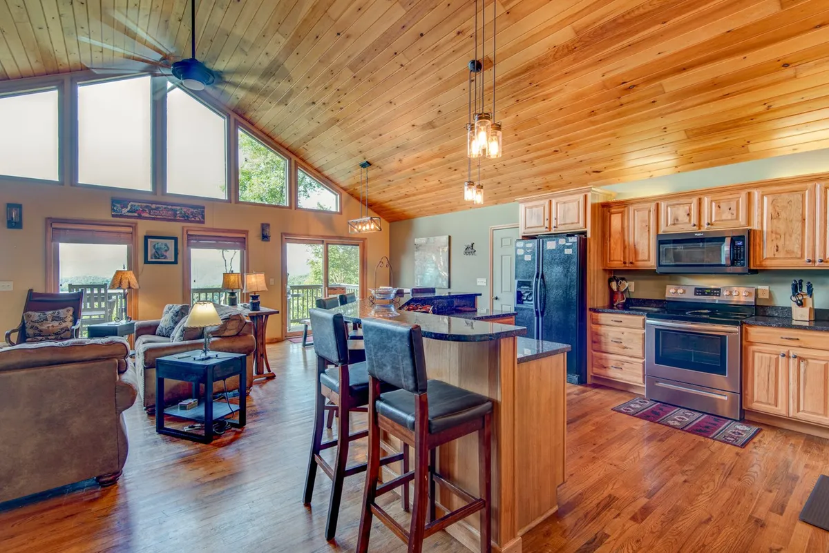 A spacious open-plan main area featuring a vaulted wooden ceiling, a kitchen island with bar stools, a comfortable seating area, and large windows offering panoramic views. The scene includes a blend of kitchen, dining, and living spaces, with warm ambient lighting and inviting furniture.