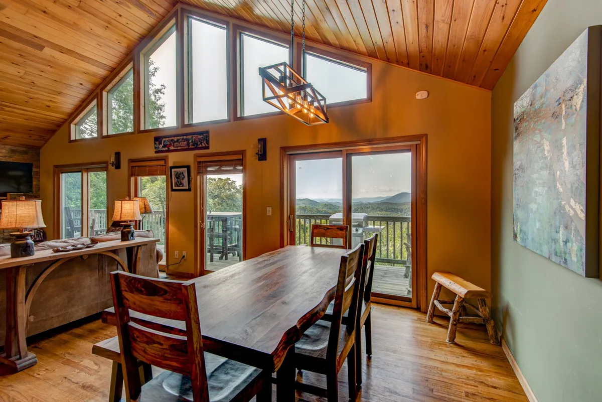 A large dining table set near panoramic windows with wooden beams overhead and artwork on the wall. The space feels bright, inviting, and ideal for gatherings and shared meals with a view.