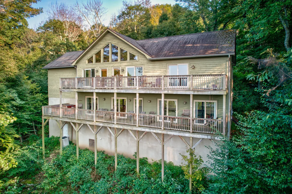The image shows a stunning mountain cabin exterior surrounded by tall trees, featuring an expansive deck and large windows that showcase breathtaking views. The deck invites guests to relax, with the promise of sunrise vistas and opportunities for stargazing in the evening, all set in a tranquil, forested environment.