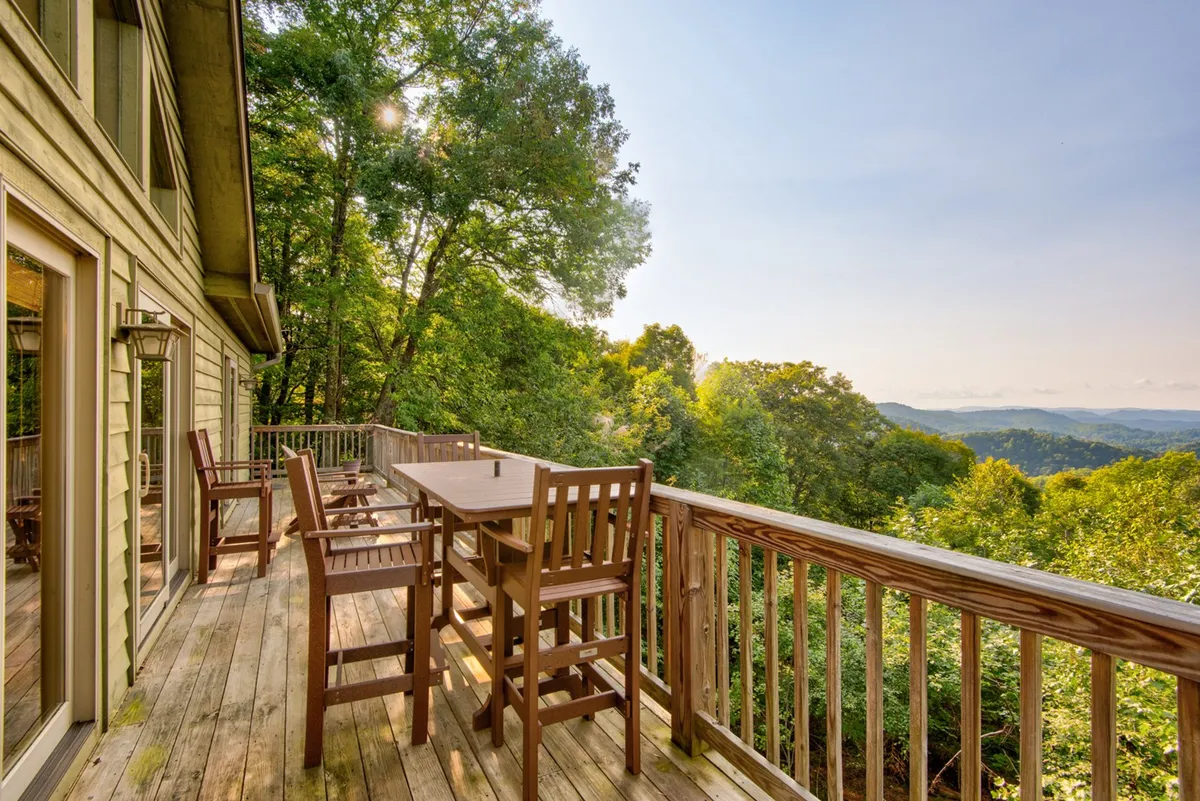 A wooden deck with bar-height table overlooks expansive mountain vistas and is surrounded by trees, offering a peaceful spot for dining, morning coffee, or sunset gatherings. The setting embodies a seamless blend of indoor comforts and outdoor enjoyment.