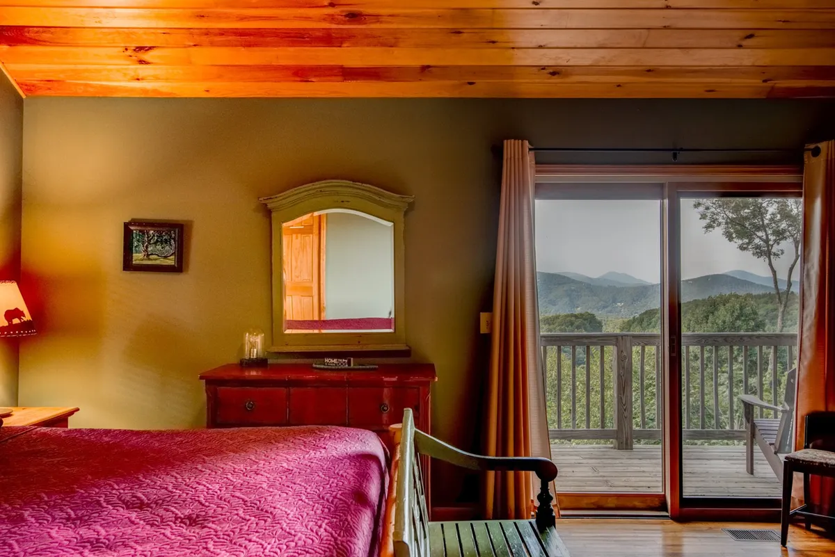 A cozy bedroom featuring a bed with a deep red quilt, a large window offering a mountain view, a wooden dresser with a mirror, and a table lamp casting a warm glow. The tranquil setup makes it ideal for relaxation and soaking in the peaceful surroundings after a day of adventure.