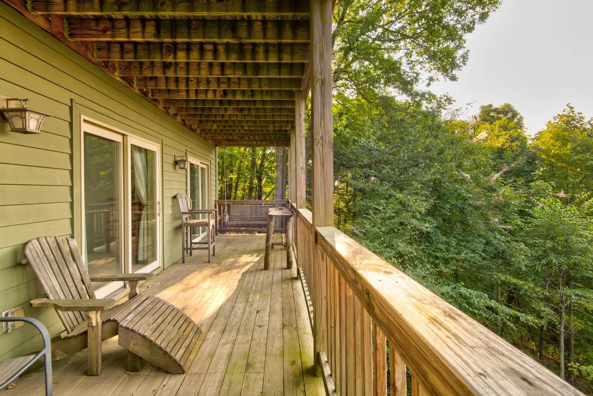 A wooden deck with comfortable Adirondack chairs overlooks the surrounding trees and features sliding glass doors that connect the interior to the outdoors. The calm, inviting scene suggests a space ideal for relaxing, sipping coffee, or gathering with friends while taking in fresh mountain air.