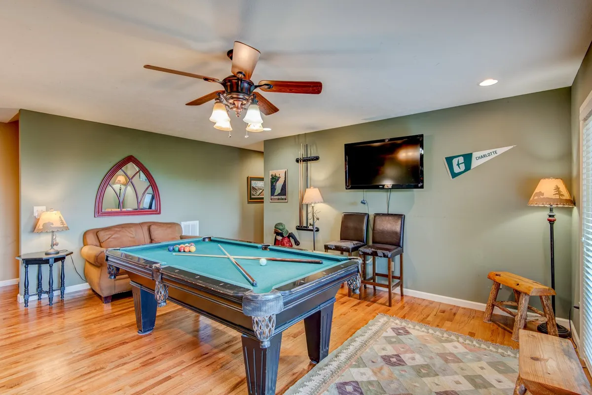 A cozy lower-level living area featuring a full-size pool table, a leather couch, a ceiling fan, a flat-screen TV mounted on the wall, and decorative wall art. The space is designed for casual fun and relaxation, inviting guests to gather for movie nights, friendly pool tournaments, or just to unwind.